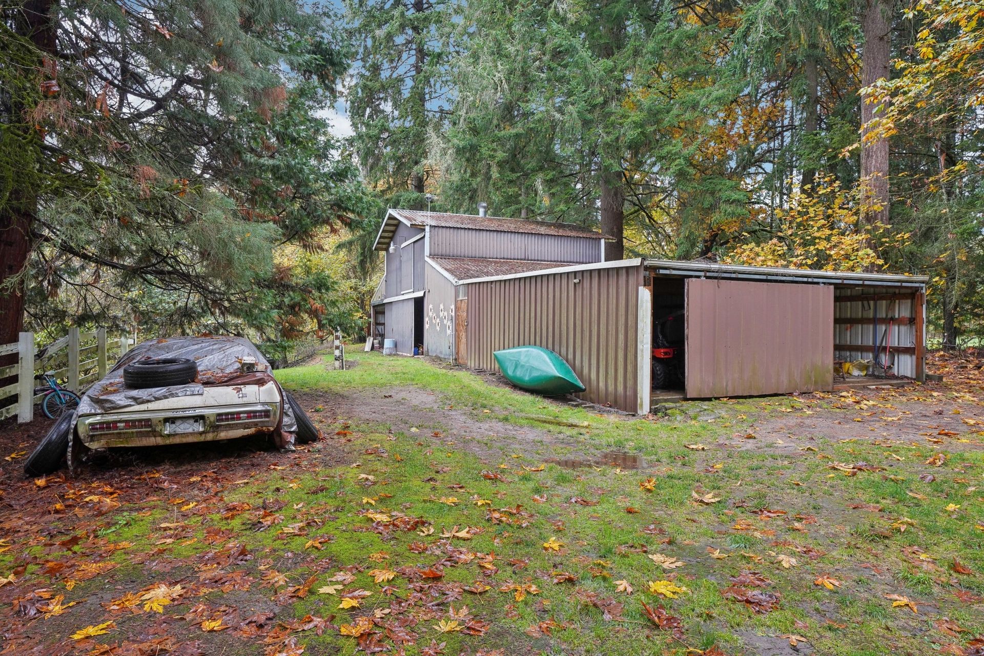 Abandoned car and rustic barn surrounded by lush forest in late autumn.